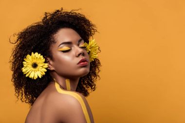 Young bright african american woman with artistic make-up and gerbera in hair dreaming isolated on orange background clipart
