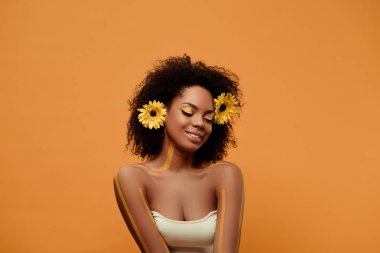 Genç gülümseyen Afrikalı-Amerikalı kadın sanatsal makyaj ve saç üzerinde turuncu arka plan izole gerbera