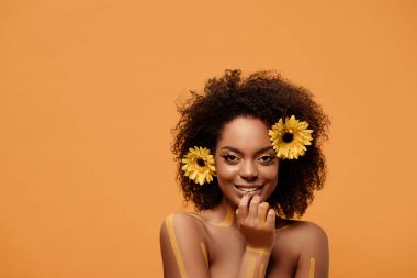 Genç gülümseyen Afrikalı-Amerikalı kadın sanatsal makyaj ve saç üzerinde turuncu arka plan izole gerbera