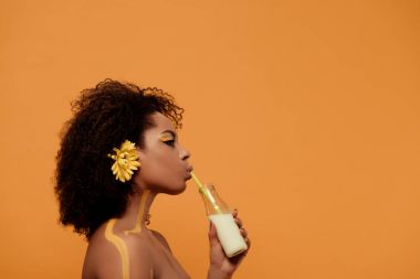 Young sensual african american woman with artistic make-up and gerbera in hair drinking milk from bottle isolated on orange background clipart