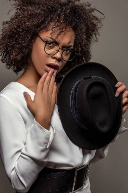 Young perplexed african american woman in white shirt wearing glasses and holding hat isolated on grey background clipart