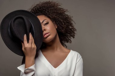 Çekici genç Afro-Amerikan kadın üzerinde izole yüzünün yarısı gri arka plan üzerinde beyaz gömlek holding siyah şapkalı