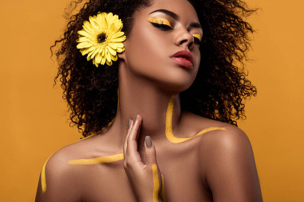 Stylish african american woman with artistic make-up and gerbera in hair tenderly touches her skin isolated on orange background