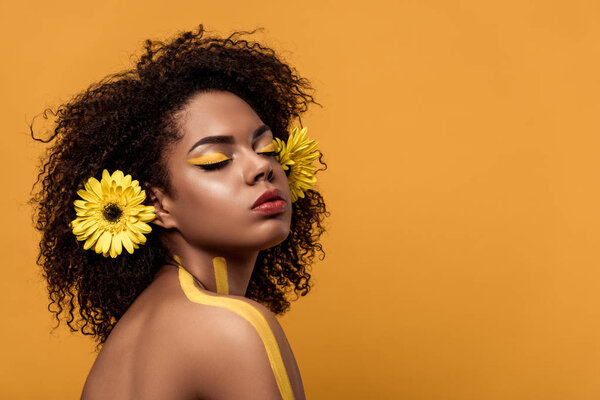 Young bright african american woman with artistic make-up and gerbera in hair dreaming isolated on orange background