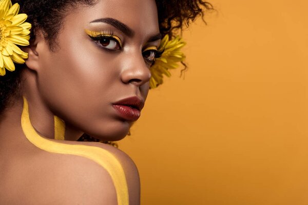 Young sensual african american woman with artistic make-up and gerbera in hair isolated on orange background