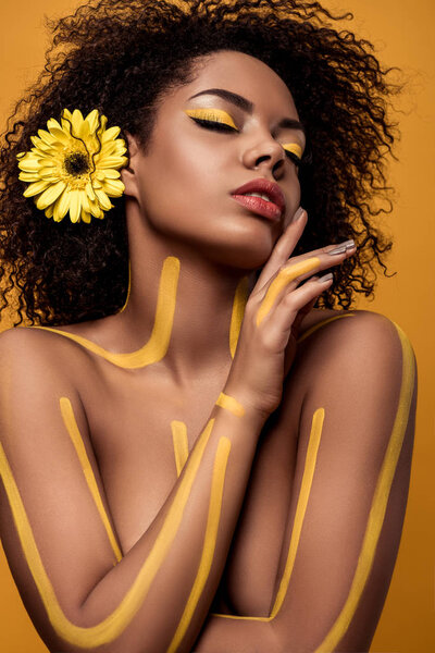 Young sensual african american woman with artistic make-up and gerbera in hair isolated on orange background