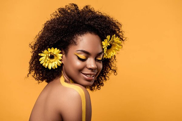 Молодая чувственная африканская американка с художественным макияжем и герберами в волосах, изолированных на оранжевом фоне
