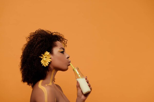 Young sensual african american woman with artistic make-up and gerbera in hair drinking milk from bottle isolated on orange background