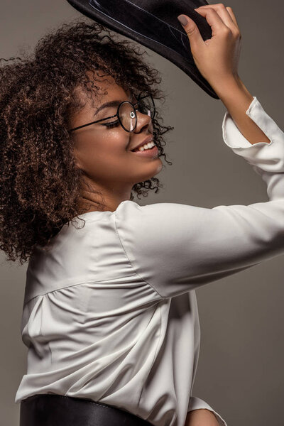 Attractive young american woman in white shirt wearing glasses pits black hat on isolated on grey background