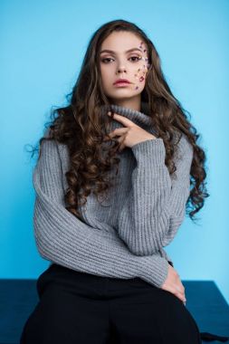 stylish long haired young woman with flowers on face in sweater sitting on table isolated on blue