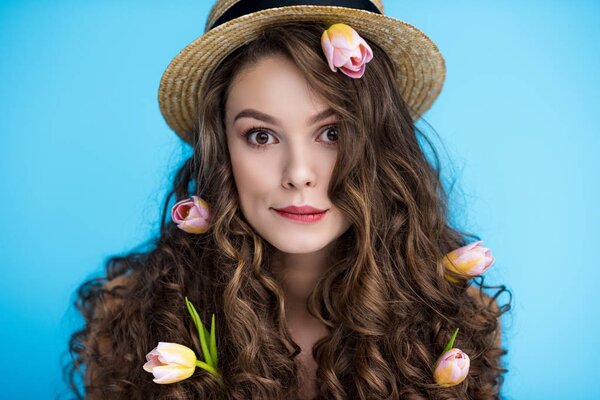 игривая молодая женщина в канонерской шляпе с цветами в длинных вьющихся волосах, смотрящая в камеру
