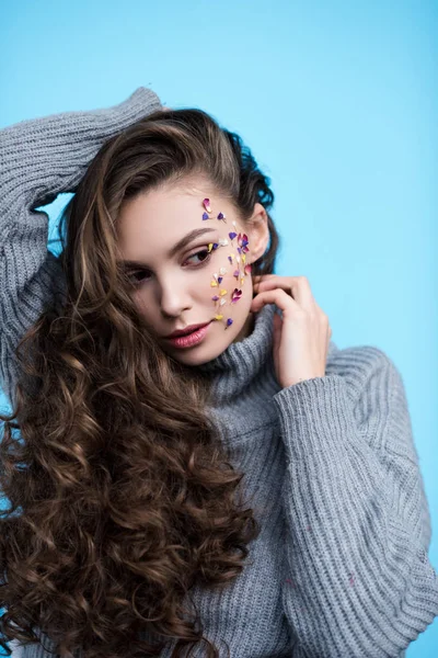 青に分離された温かみのあるニットのセーターに顔の上に花を持つ魅力的な若い女性 — ストック写真