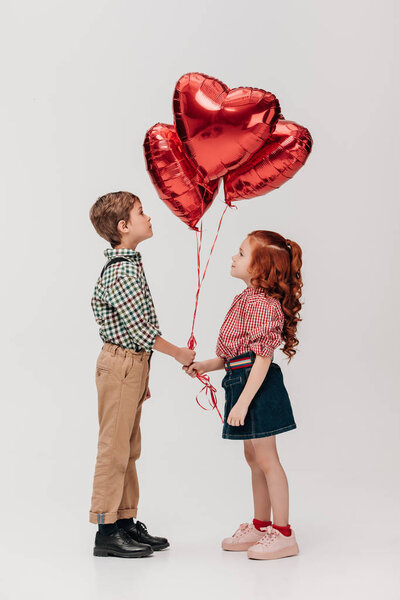 side view of beautiful little couple holding heart shaped balloons isolated on grey 