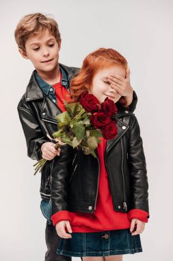 boy presenting roses bouquet to his little girlfriend and covering her eyes with hand isolated on grey clipart