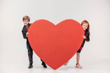 funny little kids couple hiding behind large red heart isolated on grey clipart