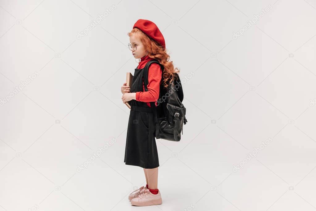 side view of adorable little schoolgirl in red beret with book and backpack isolated on grey