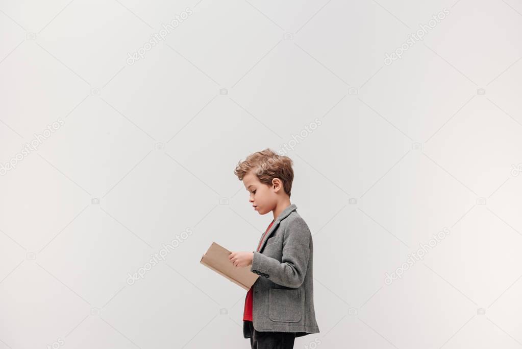 side view of stylish little schoolboy reading book isolated on grey