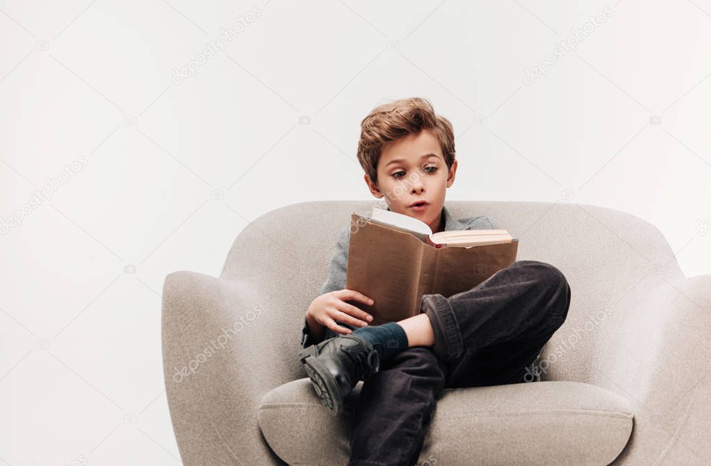 excited little schoolboy reading book in armchair isolated on grey