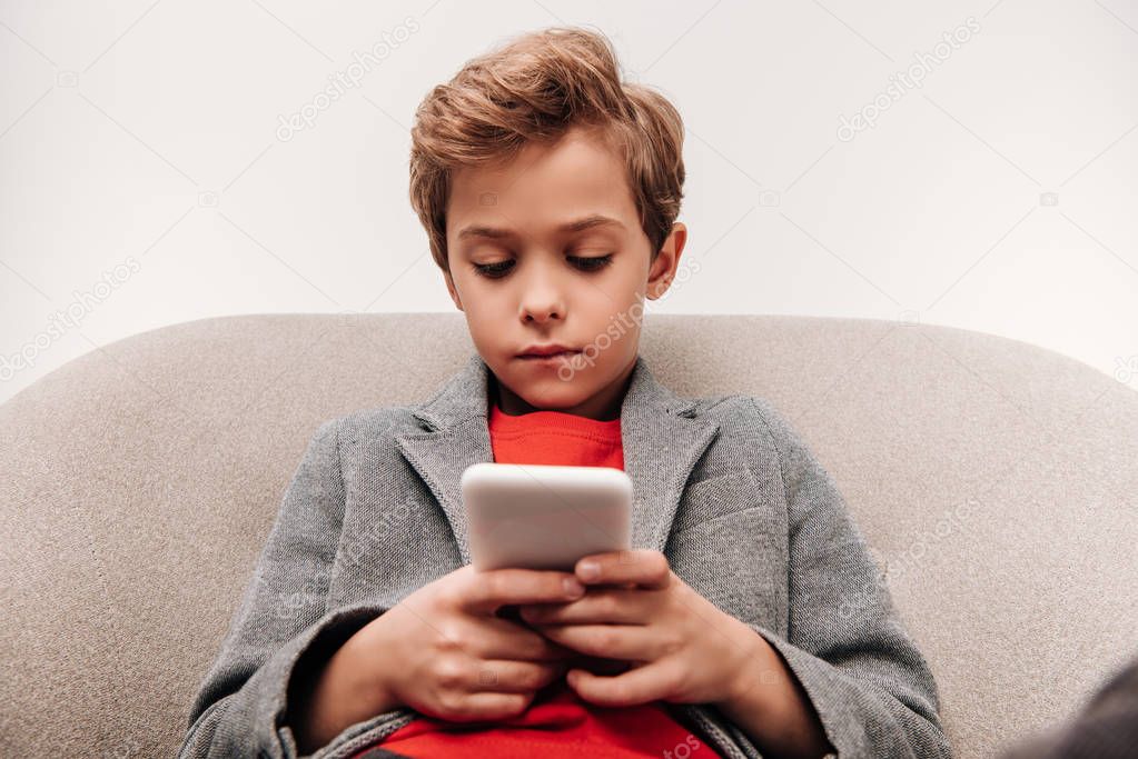 serious little boy using smartphone while sitting in armchair isolated on grey