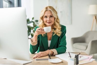 Orta yaşlı işkadını içme kahve gülümseyen ve işyerinde masaüstü bilgisayar kullanarak