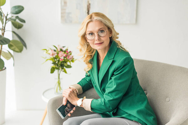 зрелая бизнесвумен в очках держит смартфон и смотрит в камеру
