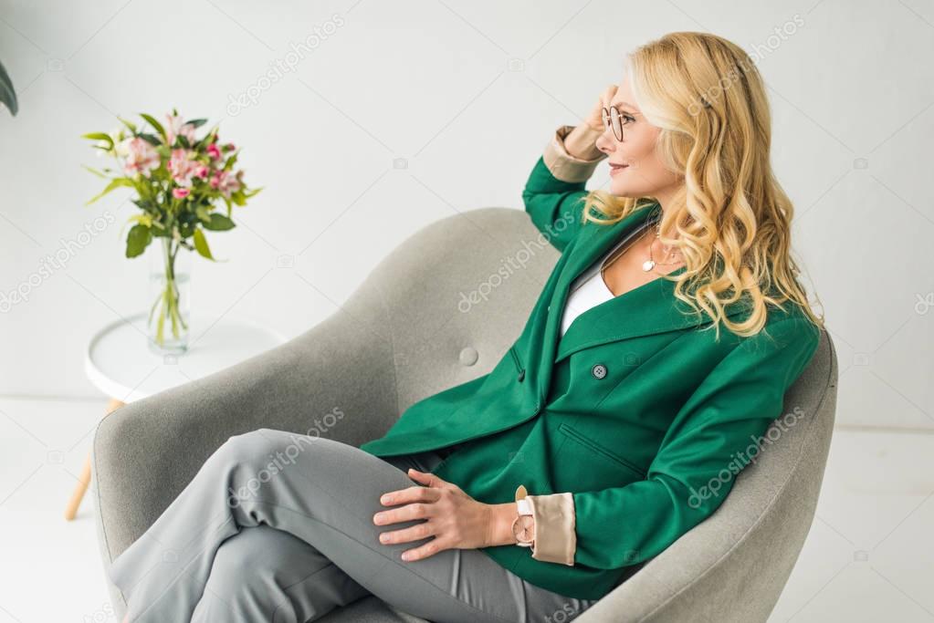 portrait of beautiful mature woman in eyeglasses sitting in armchair and looking away