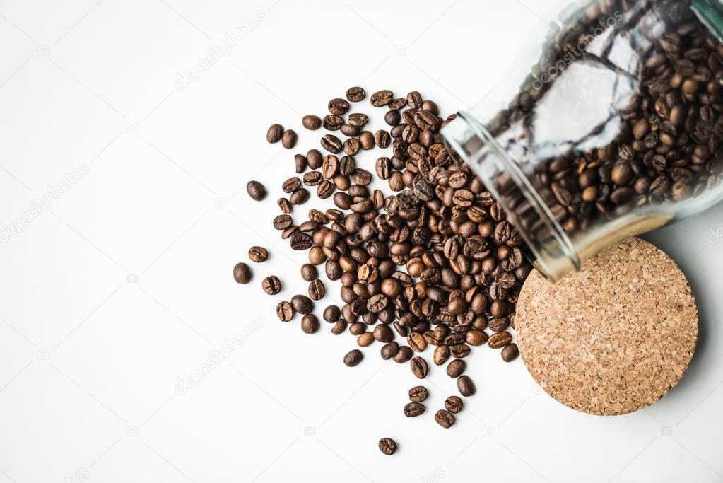 top view of scattered coffee beans from glass bottle and cork isolated on white