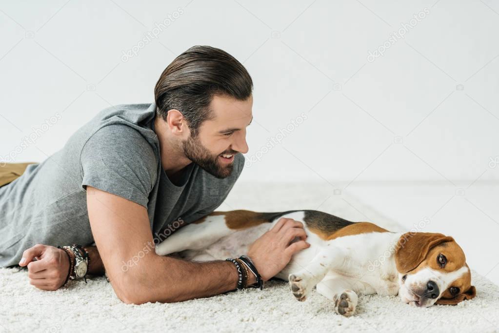 smiling handsome man lying with cute beagle on carpet