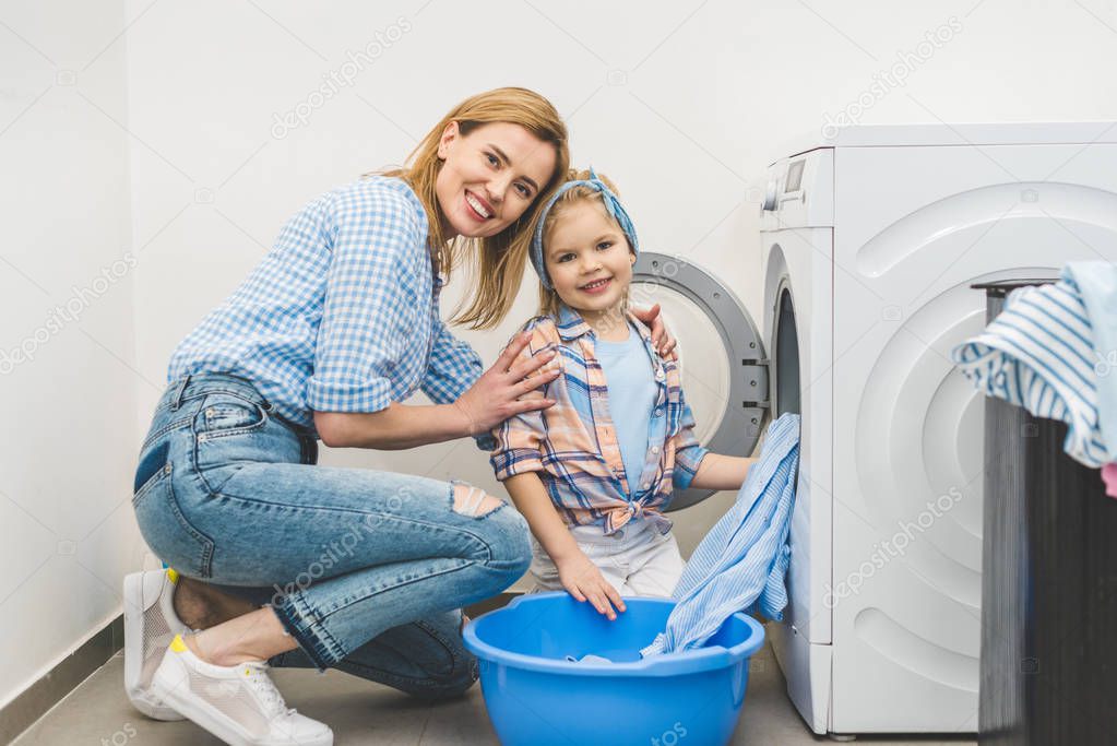 mother and daughter looking at camera while putting clothes into washing machine at home