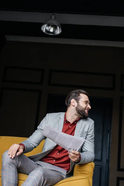 stylish man with newspaper in trendy grey suit sitting on couch in loft interior