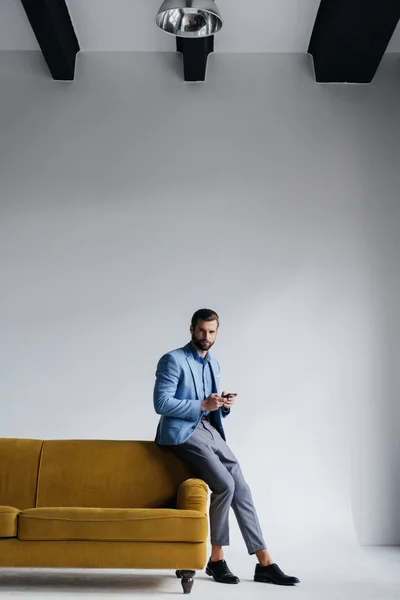 时尚男子穿着蓝色西装在黄色沙发上使用智能手机 — 图库照片