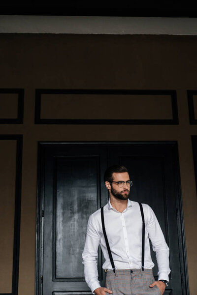 Красивый бородатый мужчина позирует в белой рубашке и подтяжки против двери
