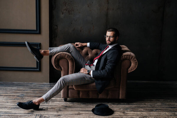 handsome elegant businessman posing in armchair, hat lying on floor near, loft interior