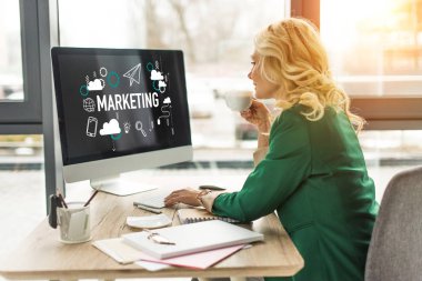 middle aged businesswoman using desktop computer with marketing icons on screen and drinking coffee at workplace  clipart