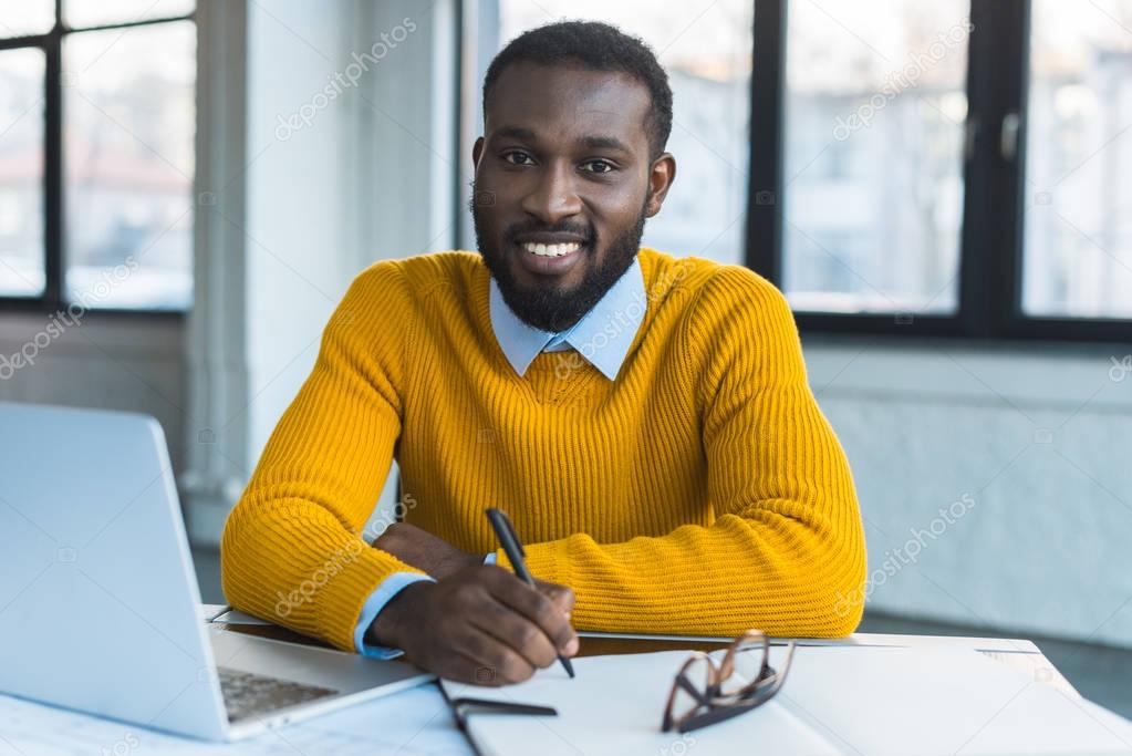 smiling african american businessman holding pen and looking at camera in office