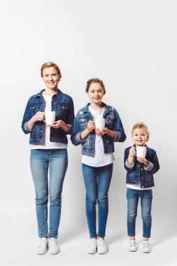 smiling mother and daughters in similar denim clothing with cups of hot drinks isolated on grey clipart