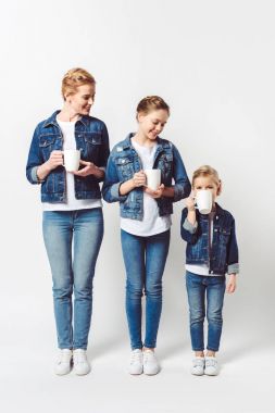 mother and daughters in similar denim clothing with cups of hot drinks standing in row isolated on grey clipart
