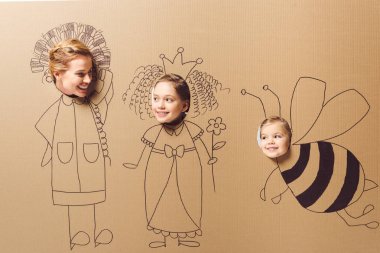 partial view of smiling mother and daughters with heads in cardboard paper with pictures clipart
