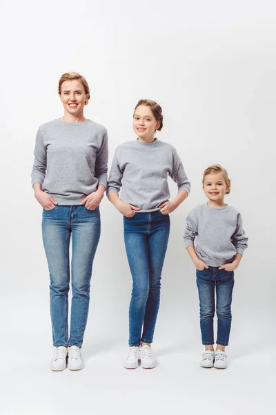 smiling mother and daughters of different generations in similar clothing with hands in pockets isolated on grey