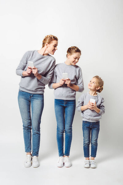 smiling mother and daughters in similar clothing with smartphones isolated on grey