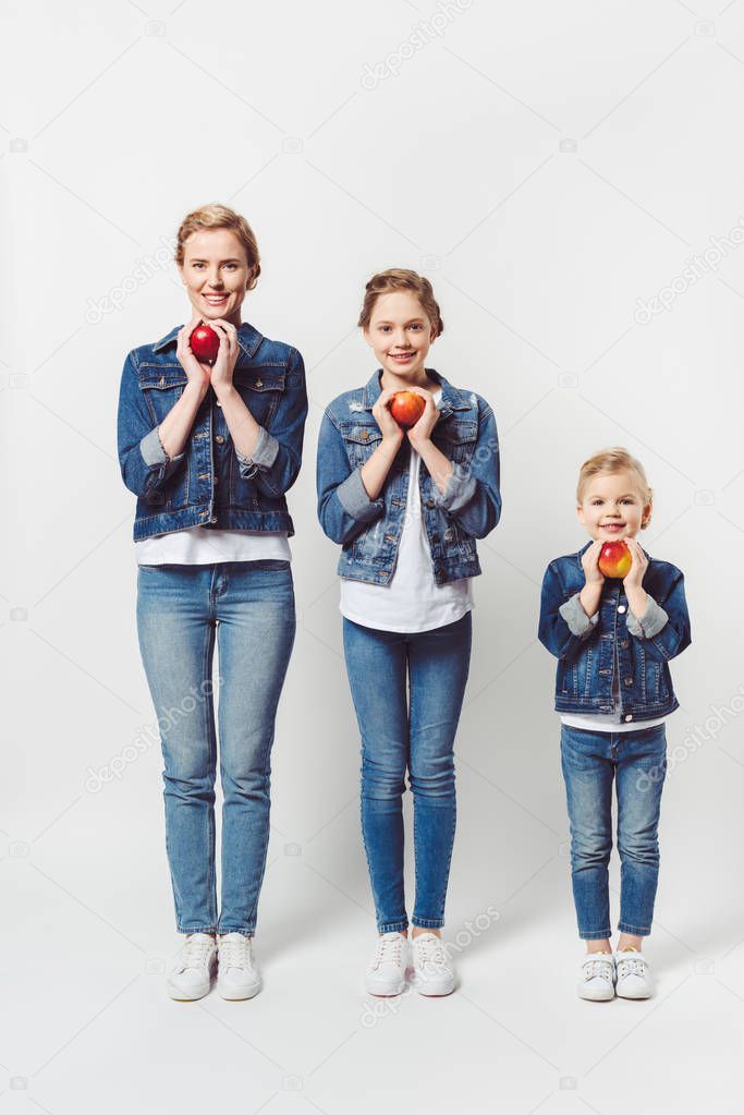smiling mother and daughters of different generations in similar clothing with fresh apples isolated on grey