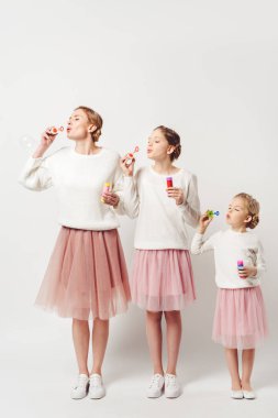 mother and daughter in similar clothing blowing soap bubbles together isolated on grey clipart