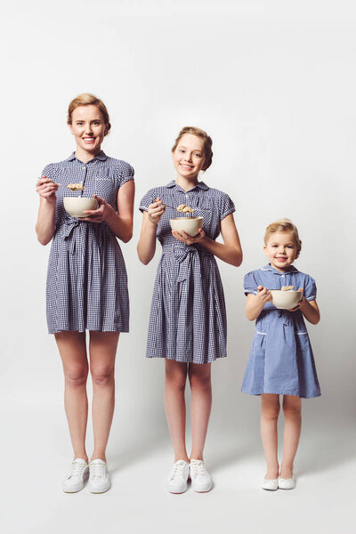 mother and daughters in similar dresses with cereal breakfast in bowls on white
