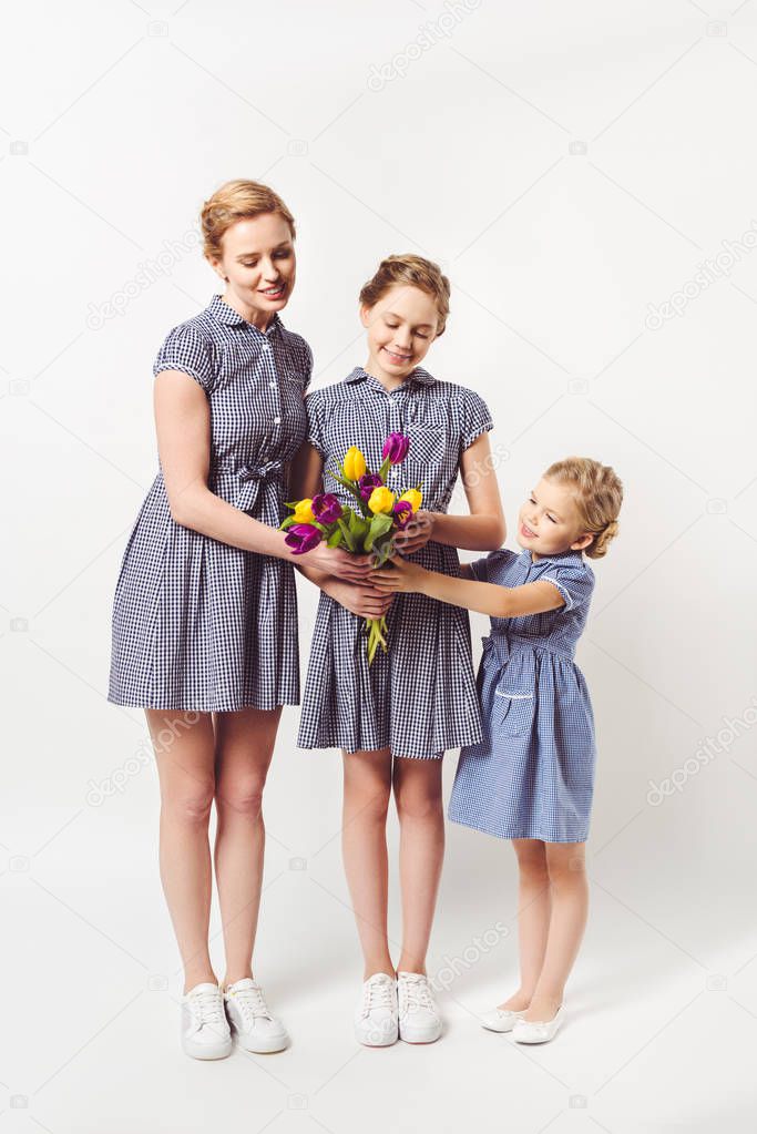 smiling mother and daughters in similar dresses with bouquet of tulips isolated on grey