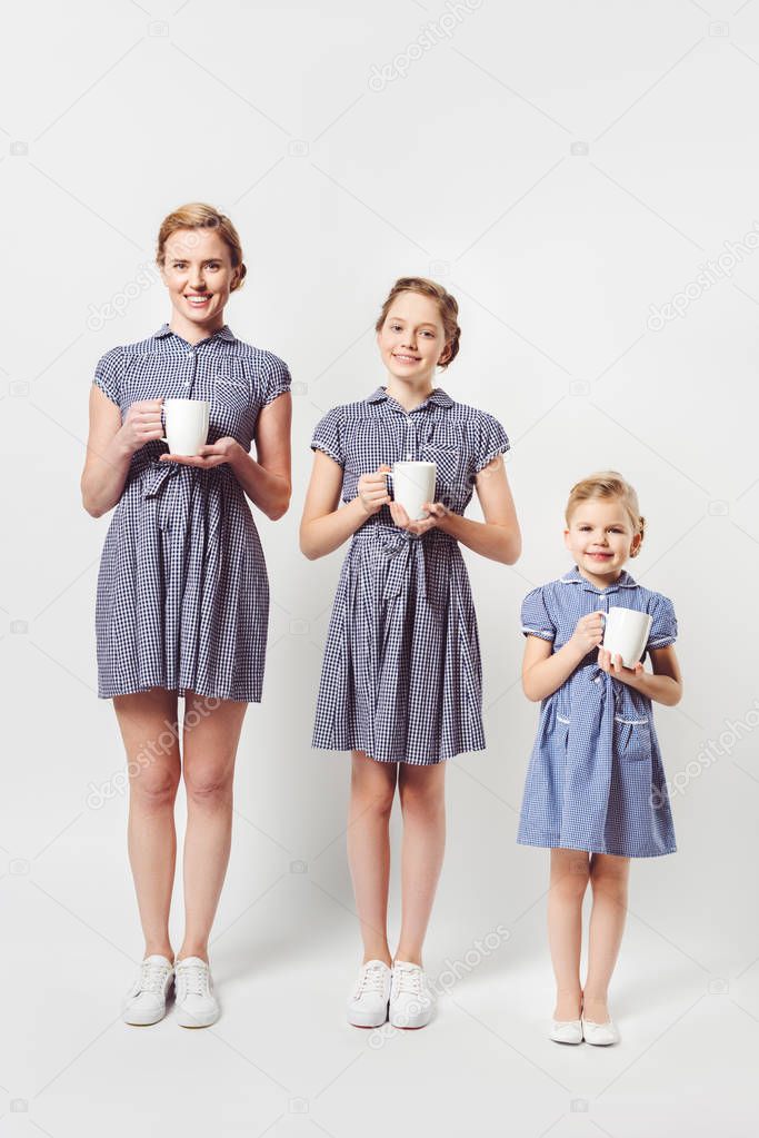 smiling mother and daughters in similar dresses with cups in hands isolated on grey