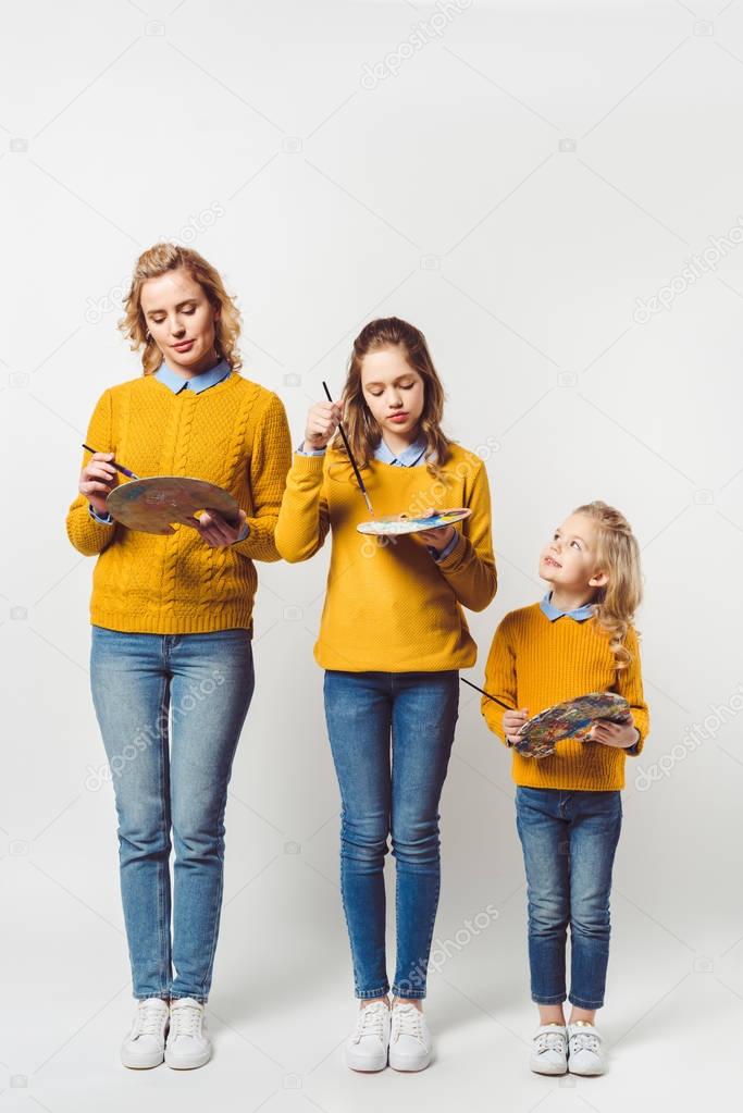 artistic mother and daughters with paint brushes and palettes on white