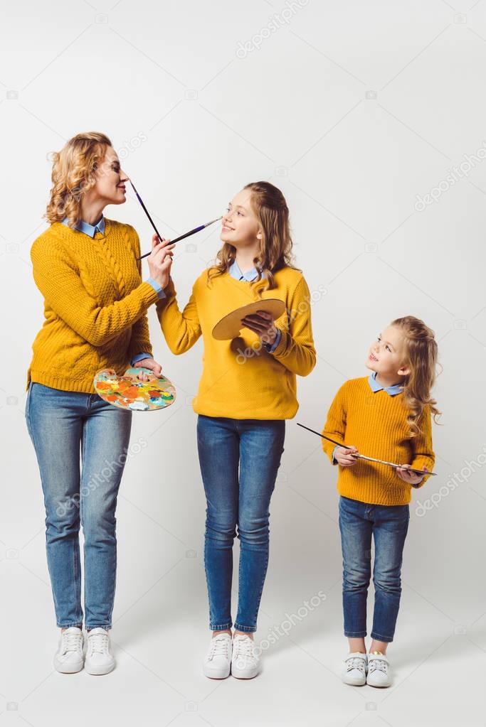 playful mother and daughters with paint brushes and palettes on white