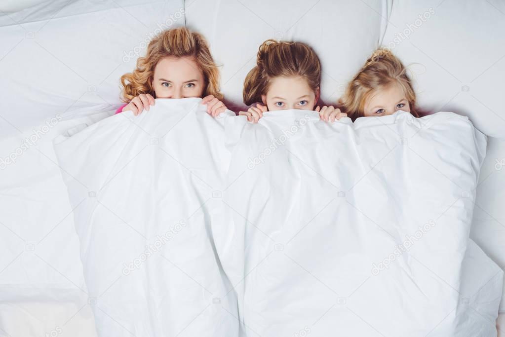 top view of mother and daughters hiding under blanket in bed