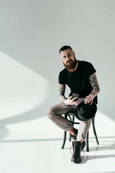 Pohledný Vousatý Tetovaný Muž Sedí Židli Smartphone Klobouk Bílém — Stock fotografie