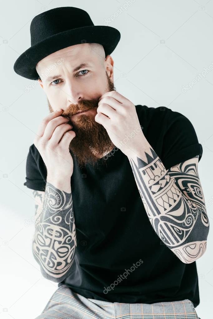 bearded tattooed man twisting mustache and looking at camera isolated on white
